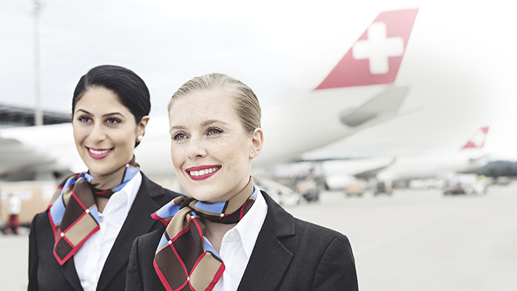 Two smiling SWISS cabin crew members on a runway, behind them several SWISS aircraft