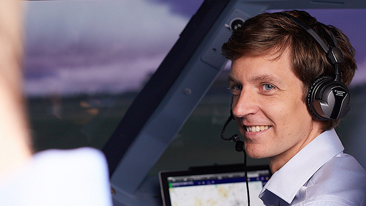 A man wearing headphones sits in an aircraft cockpit and smilingly looks at his colleague