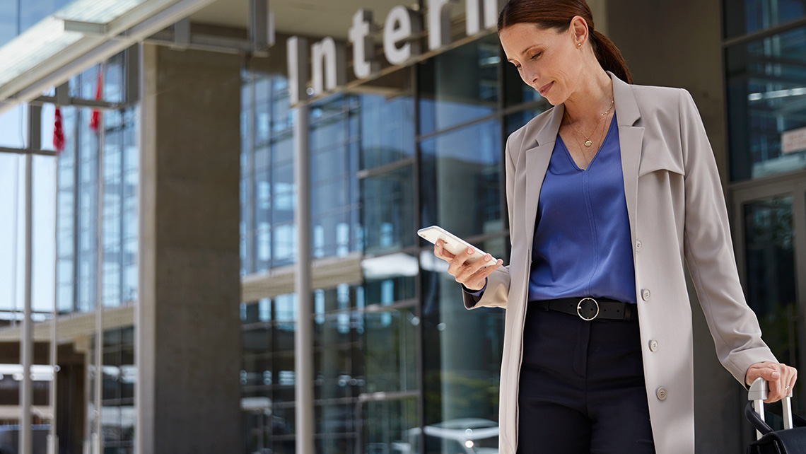 A businesswoman stands with her suitcase on wheels in front of an airport entrance and looks at her smartphone